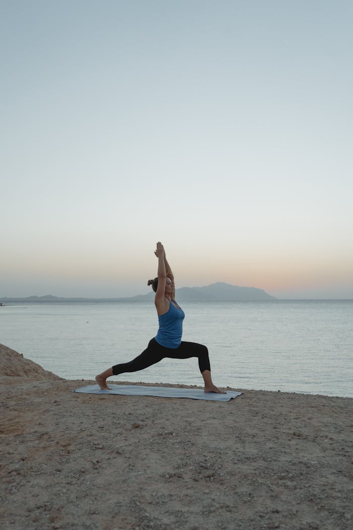A woman performs Warrior 1 yoga pose on a yoga mat at twilight by the sea, promoting tranquility and fitness.